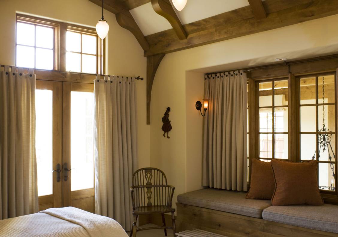 Exposed timber beams articulate the ceiling of a bedroom with an interior window seat that opens to the central space.