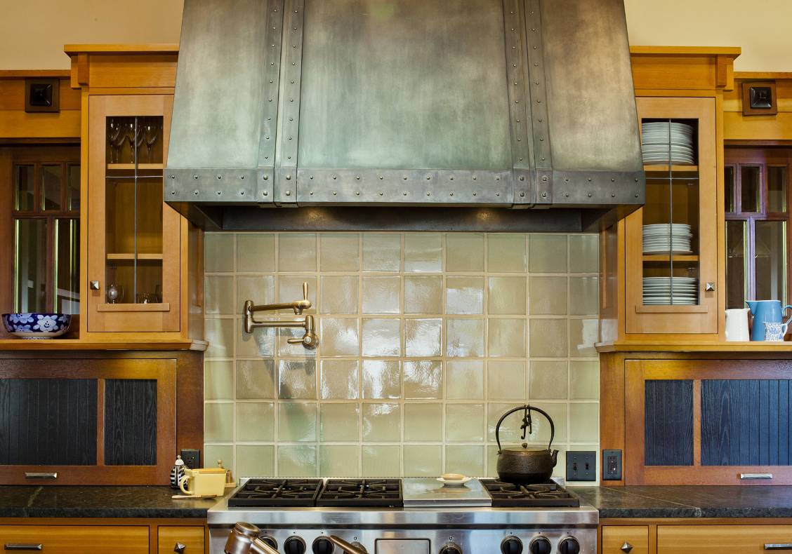 A professional stainless steel range is flanked by custom-made stained wood cabinets and black soapstone slab countertop. Hand-made ceramic tiles lined the back splash and a custom-made metal hood tops the range.