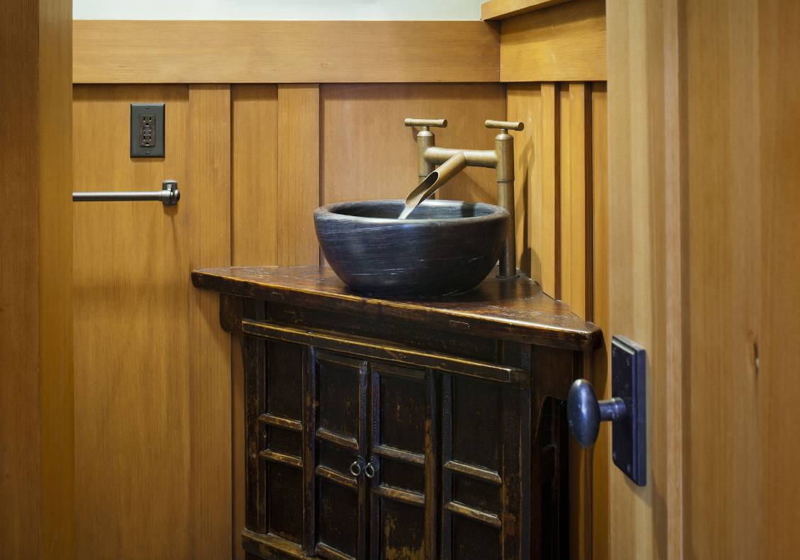 A customized antique Japanese cabinet holds a stone vessel and copper faucets. A stained wood wainscot dresses the walls.