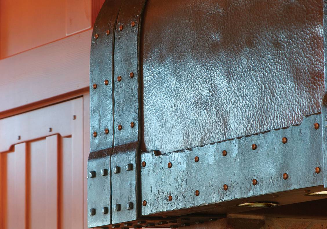 Hammered copper and iron kitchen hood with brass fasteners hovers over the range and cooking hearth, supported by reclaimed brick supports.